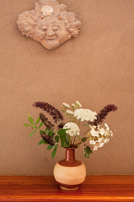Whimsical ceramic face on wall with a flower filled vase below